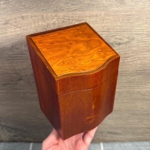 May include: A wooden box with a curved top and sides. The box is a warm brown color with a lighter, patterned wood grain on the lid. The box is held in a hand.