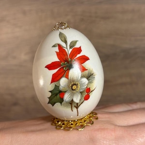 Puede incluir: Un huevo blanco con una parte superior dorada, decorado con una flor de pascua roja y una flor blanca, sobre un soporte dorado.