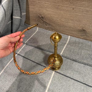 May include: A brass hookah with an ornate design. The hookah has a long, flexible hose with a colorful braided pattern. The hookah is a traditional smoking device, often used for social gatherings and relaxation.