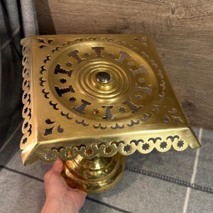 Op de afbeelding: Een messingkleurige, vierkante decoratieve standaard met een geschulpte rand en ingewikkelde uitgesneden ontwerpen. De standaard heeft een ronde basis en een centrale, donkergeleurde knop. Het algehele ontwerp is sierlijk en vintage.