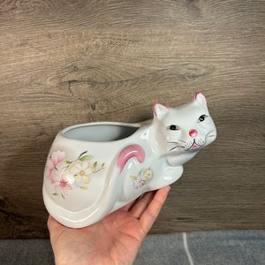 Op de afbeelding: Witte keramische kattenpot met roze oren en een bloemenontwerp. De pot is gevormd als een kat die op zijn achterpoten zit.