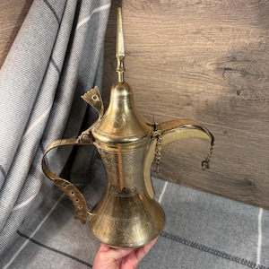 Puede incluir: Una cafetera de metal de color dorado con un diseño ornamentado y un pico largo. La cafetera tiene una tapa con un remate puntiagudo y un asa con una bisagra decorativa.