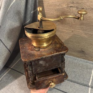 Puede incluir: Antiguo molinillo de café de madera con tapa y manivela de latón. La base de madera tiene un pequeño cajón con un pomo de latón. El molinillo está sobre un fondo a cuadros grises y blancos.
