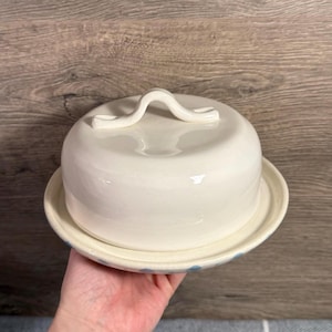 May include: A white ceramic butter dish with a domed lid and a handle. The dish sits on a matching plate with blue polka dots around the rim. The butter dish is held in a hand.