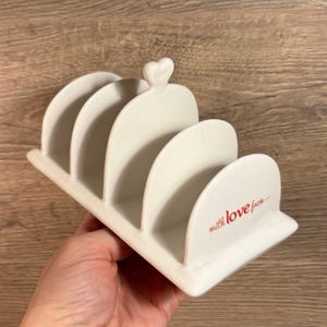 Może przedstawiać: Biały ceramiczny stojak na noże do chleba z czterema szczelinami i uchwytem w kształcie serca. Stojak ma napis "with love from..." nadrukowany na spodzie.