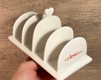 May include: White ceramic bread knife holder with four slots and a heart-shaped handle. The holder has the text "with love from..." printed on the bottom.