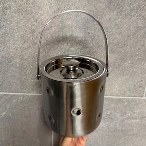 May include: A stainless steel ice bucket with a lid and a handle. The bucket has a cylindrical shape with several circular indentations. The handle arches over the top, and the lid has a small knob. The bucket is made of shiny, reflective metal.