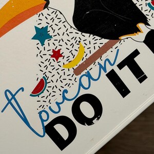 May include: A white surface featuring a graphic illustration of a toucan with a large orange beak, black body, and white chest. The design includes colorful stars, a yellow crescent moon, a watermelon slice, and the text "Toucan DO IT" in blue and black.