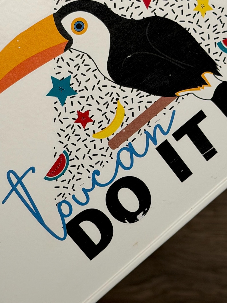 May include: A white surface featuring a graphic illustration of a toucan with a large orange beak, black body, and white chest. The design includes colorful stars, a yellow crescent moon, a watermelon slice, and the text "Toucan DO IT" in blue and black.