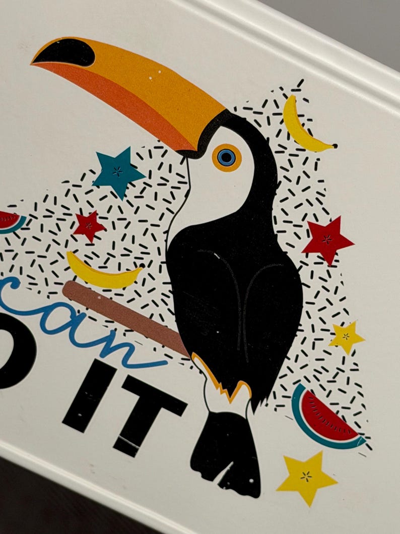 May include: A white surface featuring a graphic illustration of a toucan with a large orange beak, perched on a brown branch. The design includes yellow bananas, red and blue stars, and watermelon slices, with the partial text "can IT" in black and blue.