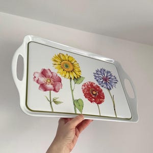 May include: White rectangular serving tray with a floral design. The tray features a sunflower, a pink rose, a red poppy, and a blue cornflower. The tray has a green border and white handles on each side.