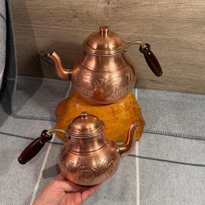 May include: Two copper teapots with rounded bodies and lids, featuring engraved circular patterns on the exterior. Each teapot has a curved spout and a dark brown wooden handle attached with brass hardware. The teapots have a metallic finish.