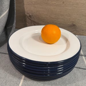 Pode incluir: Uma pilha de seis pratos redondos com um centro branco brilhante e uma borda azul-marinho escura. Uma &uacute;nica fruta laranja brilhante est&aacute; colocada no prato superior, fornecendo uma refer&ecirc;ncia visual clara para a escala do conjunto de lou&ccedil;as.