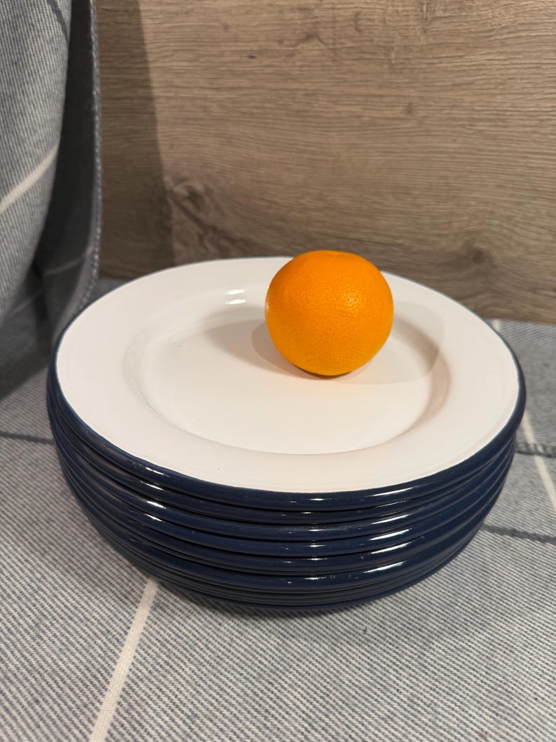 Pode incluir: Uma pilha de seis pratos redondos com um centro branco brilhante e uma borda azul-marinho escura. Uma &uacute;nica fruta laranja brilhante est&aacute; colocada no prato superior, fornecendo uma refer&ecirc;ncia visual clara para a escala do conjunto de lou&ccedil;as.