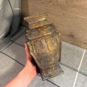 May include: A bronze-colored, antique-style vase with intricate relief carvings. The vase has a rectangular body with a flared base and a detailed rim. The carvings depict stylized figures and patterns.