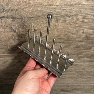 Puede incluir: Un tostador plateado con siete púas verticales y un poste central alto rematado con una pequeña bola. El soporte está hecho de metal brillante y tiene una base rectangular con un borde festoneado.