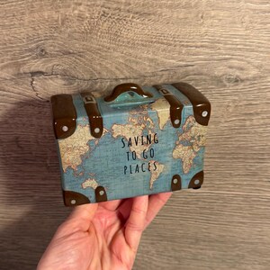 May include: A ceramic coin bank shaped like a vintage suitcase with a blue world map print, brown corner accents, and a top handle. The text SAVING TO GO PLACES is printed in black on the front. It features a coin slot on the top edge.