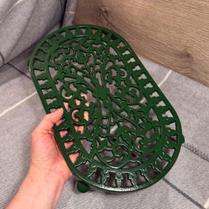 May include: An oval-shaped dark green trivet featuring an intricate, symmetrical openwork pattern of scrolling leaves and vines. The metal piece has a glossy finish and is designed with a raised border and small feet for stability on flat surfaces.
