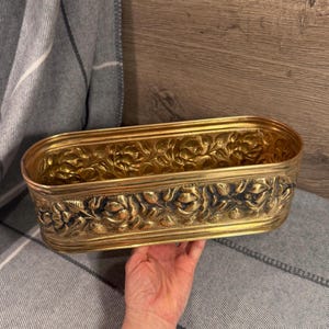 Puede incluir: Una jardinera rectangular de latón con detalles de relieve floral. La jardinera tiene forma ovalada y está sostenida por una mano. El latón tiene un tono dorado y el diseño floral está en relieve, creando una superficie texturizada.