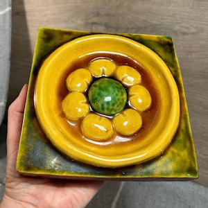 May include: A yellow and green ceramic tile with a raised design of a flower with a green center. The tile has a glossy finish.