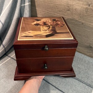 May include: A dark brown wooden jewelry box with two drawers and a decorative floral print on the lid. The box has ornate metal knobs and a rectangular base. The lid features a print of flowers and the text "LA FEE AUX ROSES".