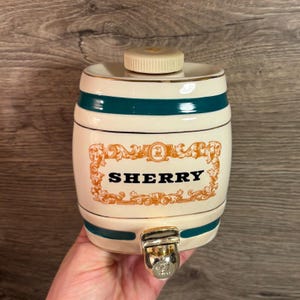 May include: Cream-colored ceramic sherry barrel with a gold-colored spigot. The barrel has a teal stripe and the word "SHERRY" in bold black letters. The barrel has a decorative gold-colored frame around the word "SHERRY".
