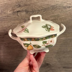May include: A white ceramic lidded serving dish with a handle, two side handles, and three feet. The dish is decorated with floral and fruit designs in pink, green, and yellow. The rim features a woven pattern.