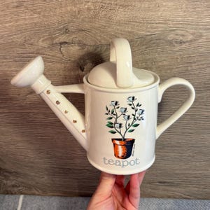 Puede incluir: Tetera de cerámica color crema con forma de regadera. Presenta un diseño floral con flores azules y hojas verdes en una maceta de terracota. La palabra "teapot" está impresa en la parte delantera.