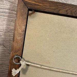 May include: A wooden picture frame with a dark brown finish. The back of the frame is visible, showing a light brown backing and a metal ring for hanging. A beige rope is attached for hanging.