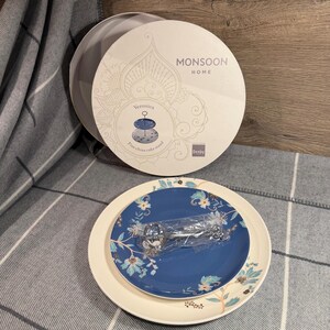Puede incluir: Un juego de soporte para pasteles de porcelana fina, con una caja redonda con el texto "MONSOON HOME" y "Veronica". El juego incluye dos platos, uno azul con motivos florales y otro color crema, y un utensilio para servir.