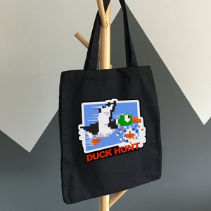 May include: Black tote bag with a white and blue pixelated image of a duck and a dog with the text "Duck Hunt" in red.