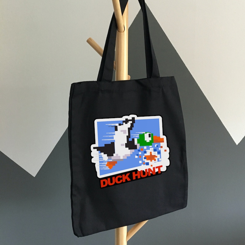 May include: Black tote bag with a white and blue pixelated image of a duck and a dog with the text "Duck Hunt" in red.
