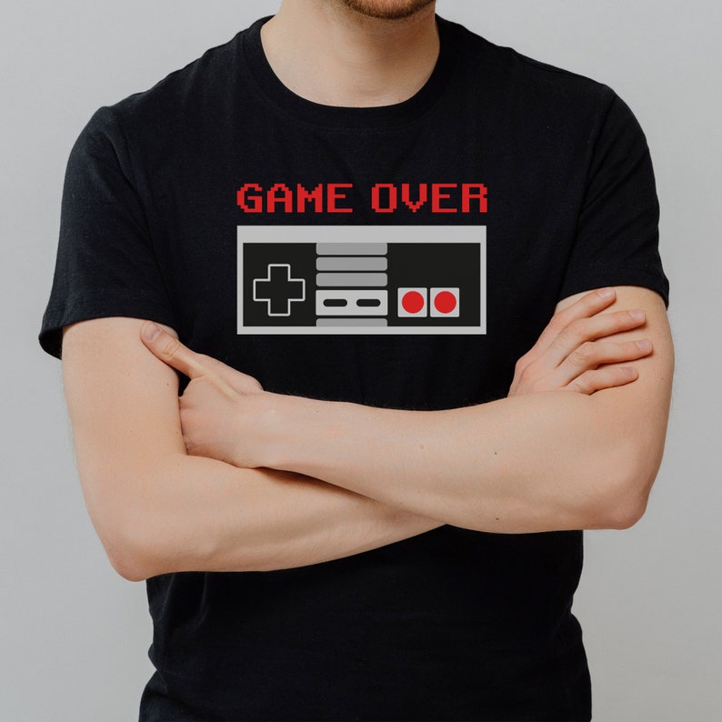 May include: A black t-shirt with the text "GAME OVER" in red pixelated letters and a pixelated image of a grey video game controller with red buttons.