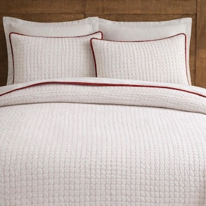 Op de afbeelding: Witte gewatteerde beddengoedset met rode bies. De set bevat een quilt en twee kussenslopen. De quilt en kussenslopen hebben een gestikt rasterpatroon. Het beddengoed wordt weergegeven op een houten hoofdbord.