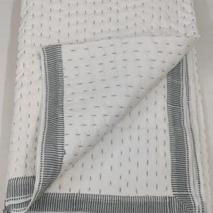 Op de afbeelding: Witte quilt met een stippenpatroon en een rand van donkergrijze strepen. De quilt is opgevouwen en onthult het interieurontwerp. De stiksels zijn zichtbaar over de hele quilt en geven het beddengoed textuur en details.