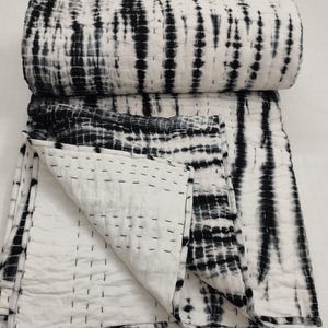 May include: A folded, tie-dye style quilt in black and white. The quilt features a repeating pattern of vertical black stripes against a white background. The edges are finished with black stitching, and the quilt is folded to display its interior.