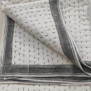 Puede incluir: Colcha blanca con detalles de costura negros y un borde a rayas. La colcha está doblada, mostrando la textura y el diseño. La costura crea un patrón sutil en la superficie, lo que aumenta el atractivo visual. La estética general es minimalista y moderna.