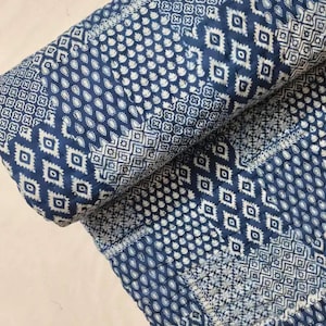 Puede incluir: Edredón enrollado con estampado azul índigo y blanco. El edredón presenta varios diseños geométricos, incluyendo diamantes, cuadrados y formas de cachemira. La tela parece ser de algodón, con un borde cosido. El diseño general es simétrico y detallado.