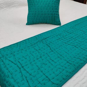 May include: A teal-colored quilted pillow and bed runner set on a white bedspread. The pillow is square, and the runner extends down the bed. The bedding has a textured, woven appearance, adding a decorative element to the bedroom.