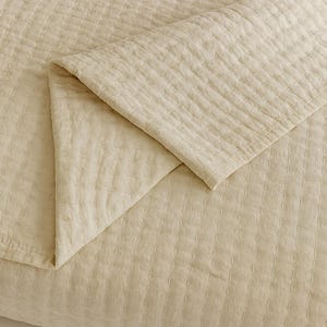 Könnte beinhalten: Eine gefaltete, beige Decke mit einem strukturierten, gesteppten Muster. Der Stoff hat einen subtilen Glanz, und die gefaltete Ecke zeigt die Dicke und Konstruktion der Decke. Die Gesamtästhetik ist schlicht und elegant, geeignet für Bettwäsche oder Heimdekoration.