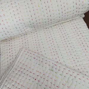 May include: White quilted blanket with colourful, evenly spaced stitching in red, yellow, and green. The blanket is folded and rolled, showcasing the detailed stitching pattern. The overall design is simple and elegant.