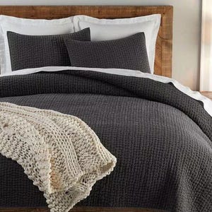 May include: A bed featuring a dark grey quilted comforter, matching pillow shams, and a cream-coloured knitted throw blanket. The bed has white sheets and a wooden headboard, creating a cosy and inviting bedroom setting.