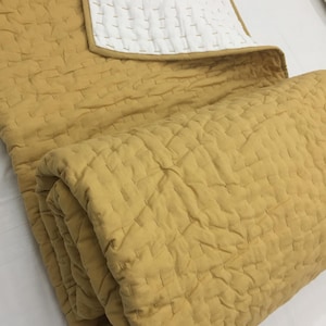 Könnte beinhalten: Senfgelbe Steppdecke mit weißer Rückseite. Die Decke hat ein gestepptes Gittermuster und einen gefalteten Bereich, der den Kontrast zwischen den beiden Seiten zeigt. Die Kanten sind mit einer kontrastierenden Paspel sauber verarbeitet.