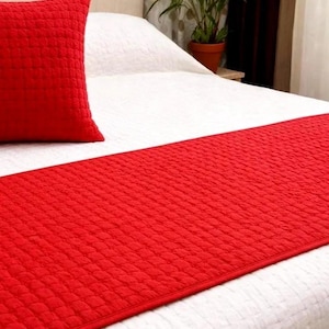 Puede incluir: Un camino de cama y una almohada a juego acolchados rojos sobre una cama blanca. El camino de cama es rectangular y cubre la parte inferior de la cama. La almohada es cuadrada y descansa sobre la cama. Una planta en maceta marrón está al fondo.