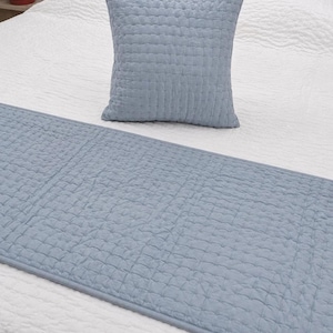 Puede incluir: Un cojín cuadrado de color azul claro acolchado y un camino de cama rectangular a juego. Ambos artículos presentan un patrón de costuras en forma de cuadrícula en toda la superficie, lo que aporta una textura al tejido azul sólido.