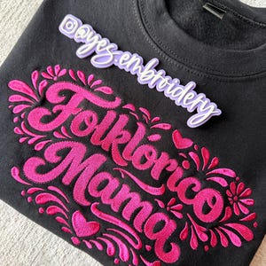 May include: Black sweatshirt with the embroidered words "Folklórico Mama" in bright pink, surrounded by floral and heart designs. The sweatshirt also has a small tag that says "@ayes.embroidery".