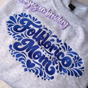 May include: A gray sweatshirt with a blue embroidered design that reads "Folklórico Mom" in a stylized script. The design is surrounded by floral and leaf-like embellishments. A purple Instagram handle is visible at the top.