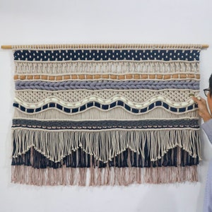 Peut inclure: Une grande tapisserie murale en macramé avec une variété de textures et de couleurs, notamment beige, marron, gris et bleu. La tapisserie a une frange en bas et est montée sur une baguette en bois.