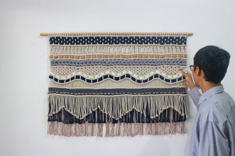 May include: A large wall hanging made of macrame with a variety of textures and colors, including beige, brown, gray, and blue. The hanging has a fringe at the bottom and is mounted on a wooden dowel.