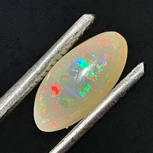 Puede incluir: Una piedra preciosa de ópalo ovalada blanca con destellos iridiscentes de arcoíris, sostenida por unas pinzas de plata.